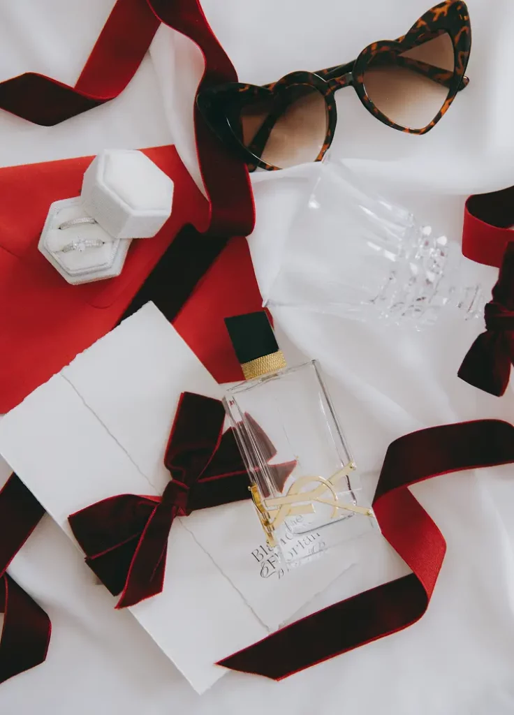 Flatlay de mariage avec rubans rouges, lunettes, alliance, parfum et papeterie pour le shooting Amour & Velour